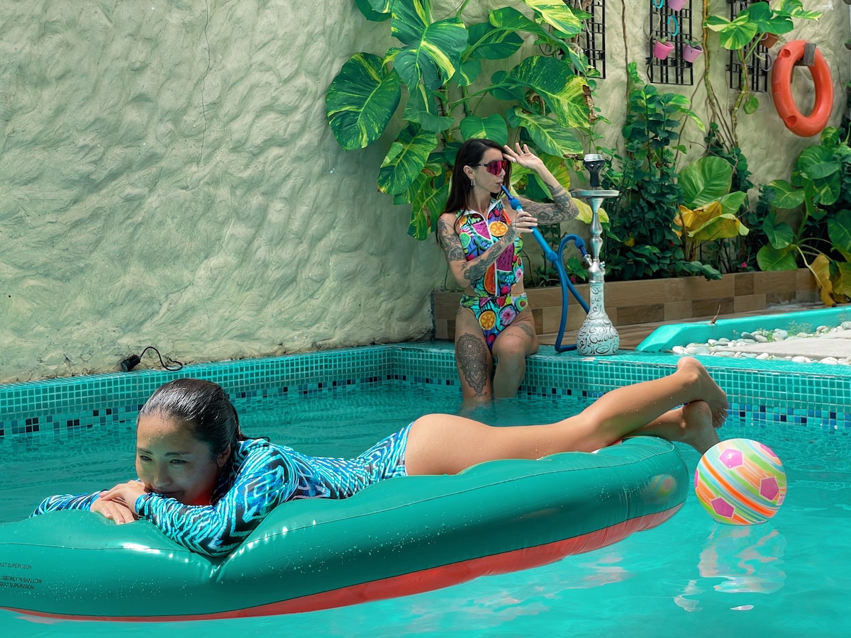A vibrant swimming pool is surrounded by lush tropical foliage. One person relaxes on a colorful inflatable float, while another is seated at the pool's edge, enjoying refreshments. A playful beach ball rests nearby, adding to the lively atmosphere.