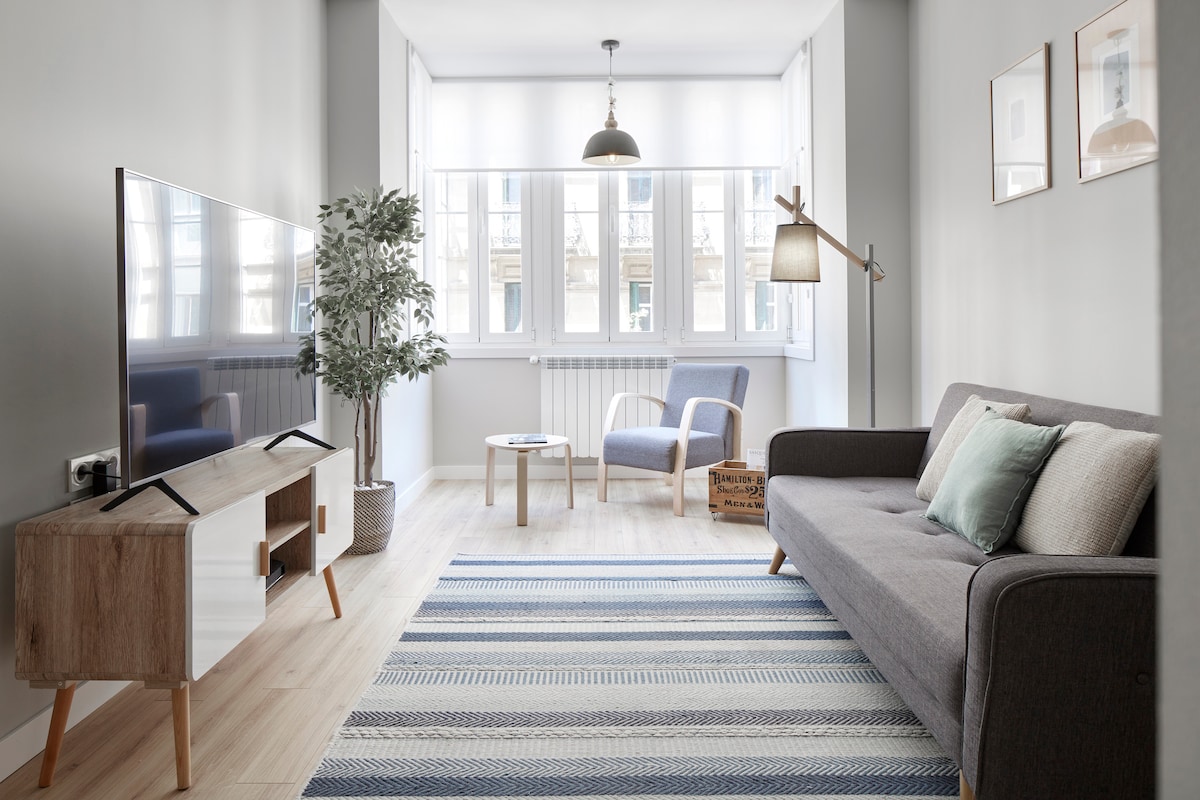A modern living area features a gray sofa and a blue armchair, complemented by a wooden coffee table and a stylish rug. Large windows allow natural light to fill the room, enhancing the serene atmosphere. A flat-screen TV is positioned on a wooden media console.