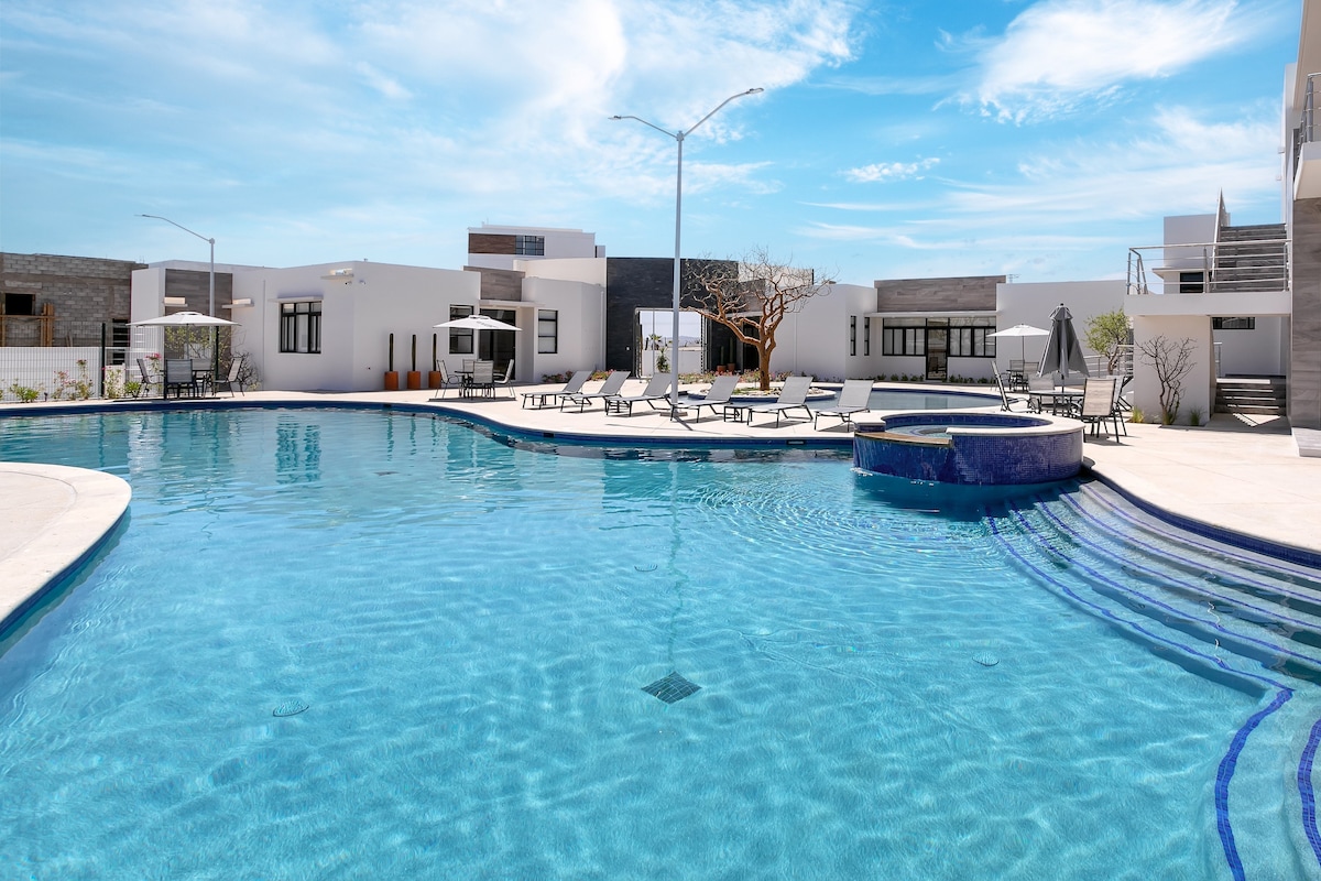 A large swimming pool is visible in the foreground, surrounded by lounge chairs and umbrellas. Modern buildings are seen in the background, with an inviting outdoor space illuminated by natural light and a clear blue sky.