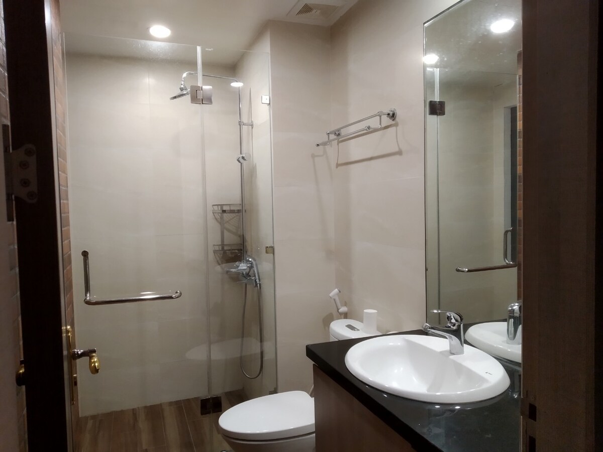 A modern bathroom features a walk-in shower with glass doors and a wall-mounted showerhead. A sleek vanity with dual sinks is complemented by a large mirror. The toilet is positioned nearby, with safety grab bars installed for accessibility. Neutral tones create a clean and spacious ambiance.