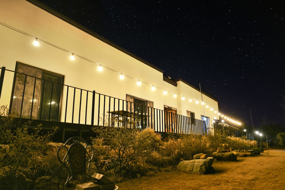 The exterior of the accommodation is illuminated by string lights, creating a gentle glow against the night sky filled with stars. A railing runs along the front, with tables and seating visible on the deck surrounded by neatly arranged rocks and low vegetation.