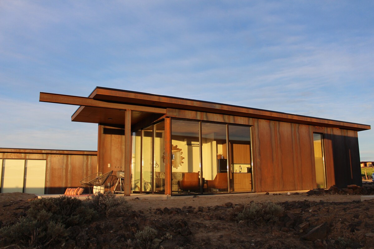A modern, southwestern-themed condo is displayed, featuring a sleek, rust-colored exterior. Large glass doors allow natural light to enter, revealing an open layout that connects the kitchen, dining, and living areas. The surrounding landscape consists of gravel and low vegetation.