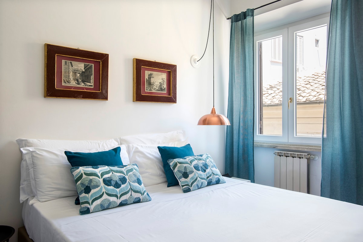 A bright bedroom is furnished with a spacious bed highlighted by patterned cushions. Above, two framed prints adorn the wall, and a pendant light adds a modern touch. Light blue curtains frame a window, offering a view of the outside.