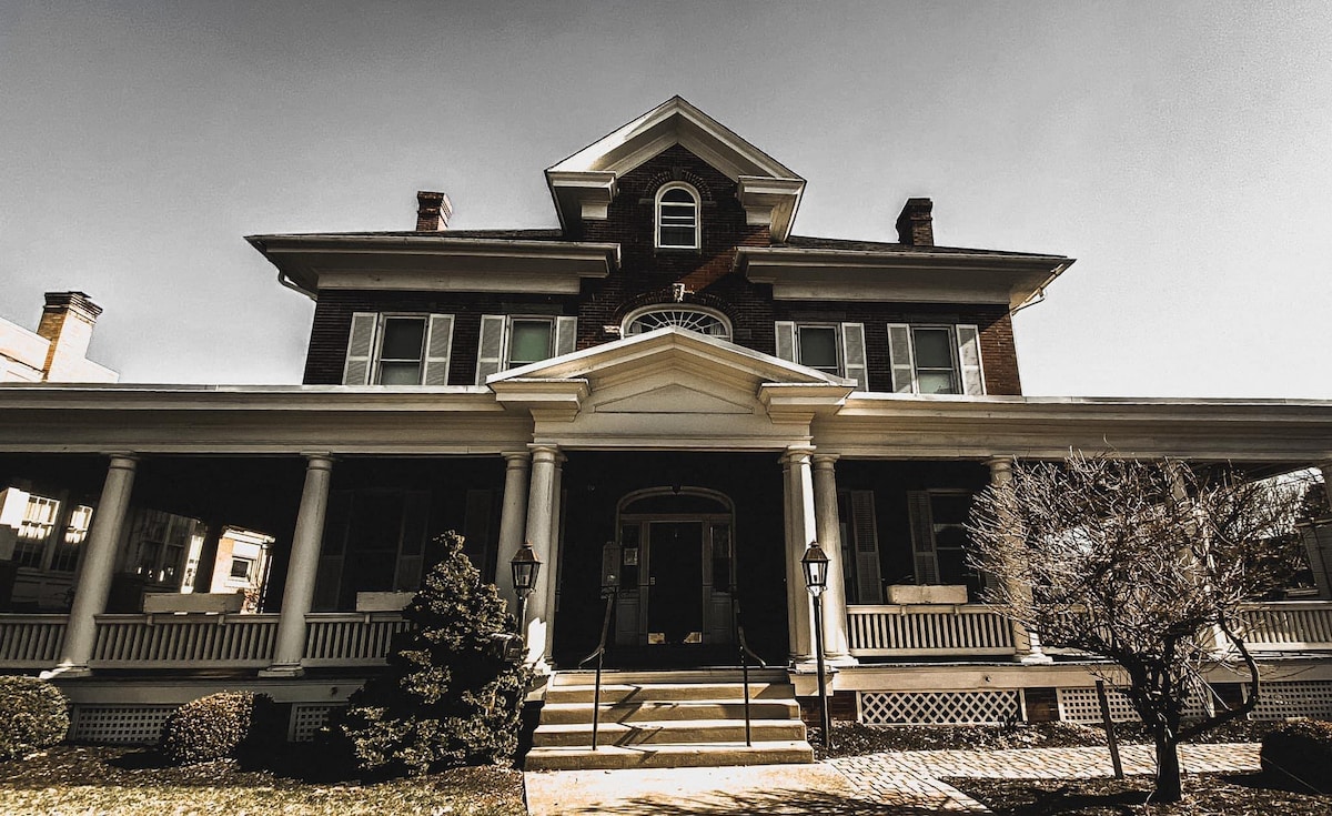 The historic 1912 Hoover House features a grand entrance with large columns supporting an expansive porch. Symmetrical windows frame the entrance, and manicured shrubs complement the front lawn. The building's brick facade reflects a classic architectural style, showcasing its prominent structure within the town.