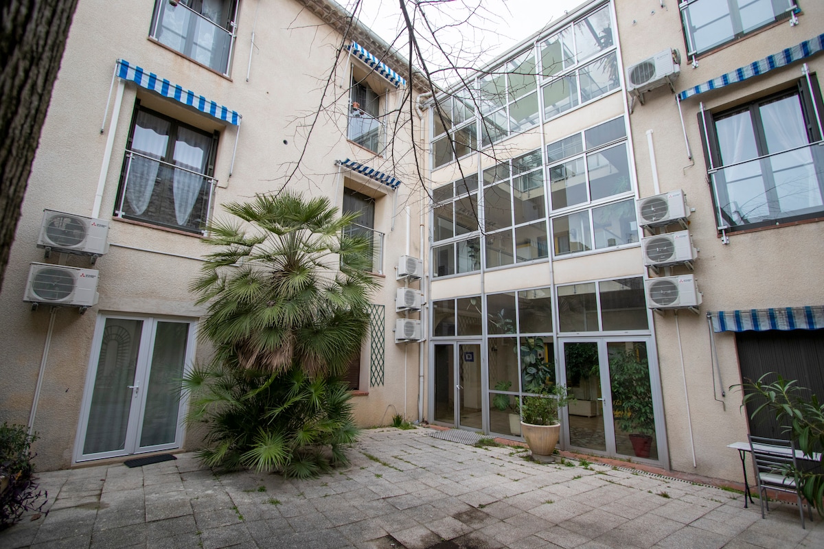 An outdoor courtyard is surrounded by three stories of the building. Air conditioning units are mounted on the exterior walls. A palm plant is positioned in a ceramic pot, complemented by a tiled floor. Large glass windows enhance the airy feel of the space.
