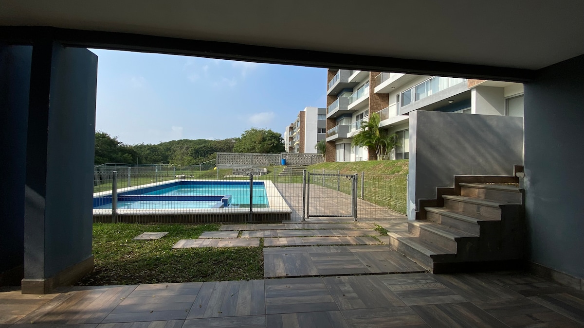 A view is presented from a covered patio overlooking a swimming pool, surrounded by green grass. Steps lead up from the patio, and residential buildings are visible in the background. A secure fence encircles the pool area, emphasizing a private outdoor space.