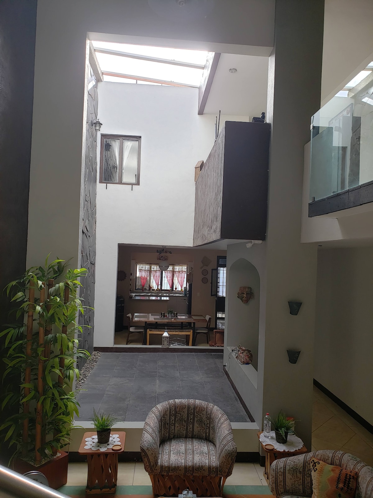 An interior view features a central open space bathed in natural light. Comfortable seating is arranged around a stone accent wall, while a dining area in the background is visible. Skylights above enhance the airy feel of the home.