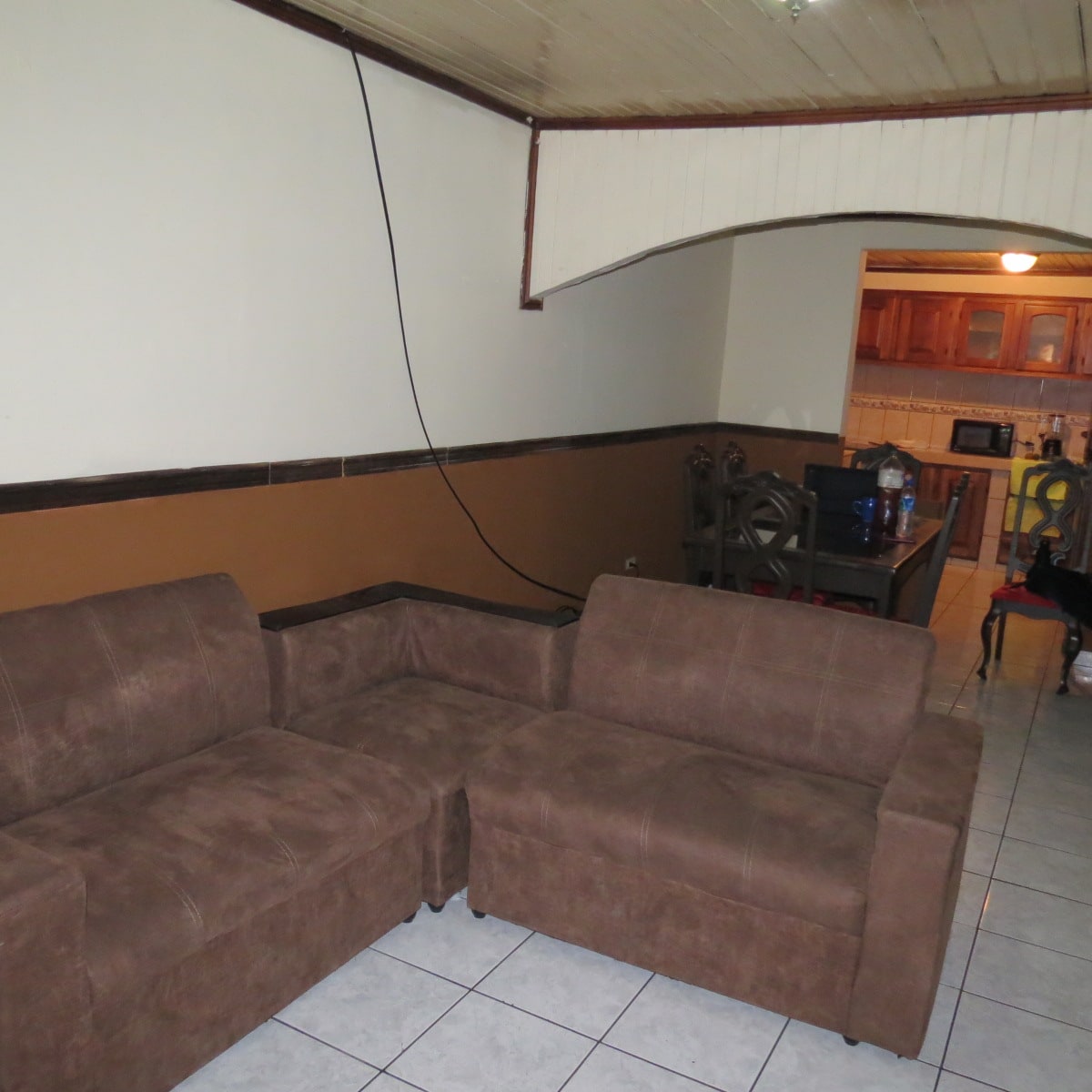 A comfortable seating area features a brown sectional sofa positioned against a neutral wall. A dining table with chairs is visible in the background, alongside an open kitchen area with wood cabinetry and appliances. Tile flooring complements the cozy environment.