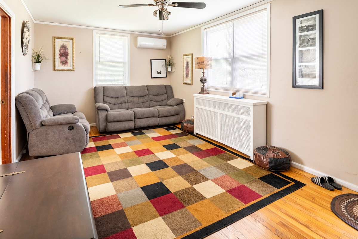 A spacious living room features a large, colorful area rug that defines the seating area. Two gray recliners are positioned near a window that allows natural light. A console table and lamp add functionality. Wooden flooring complements the cozy ambiance.