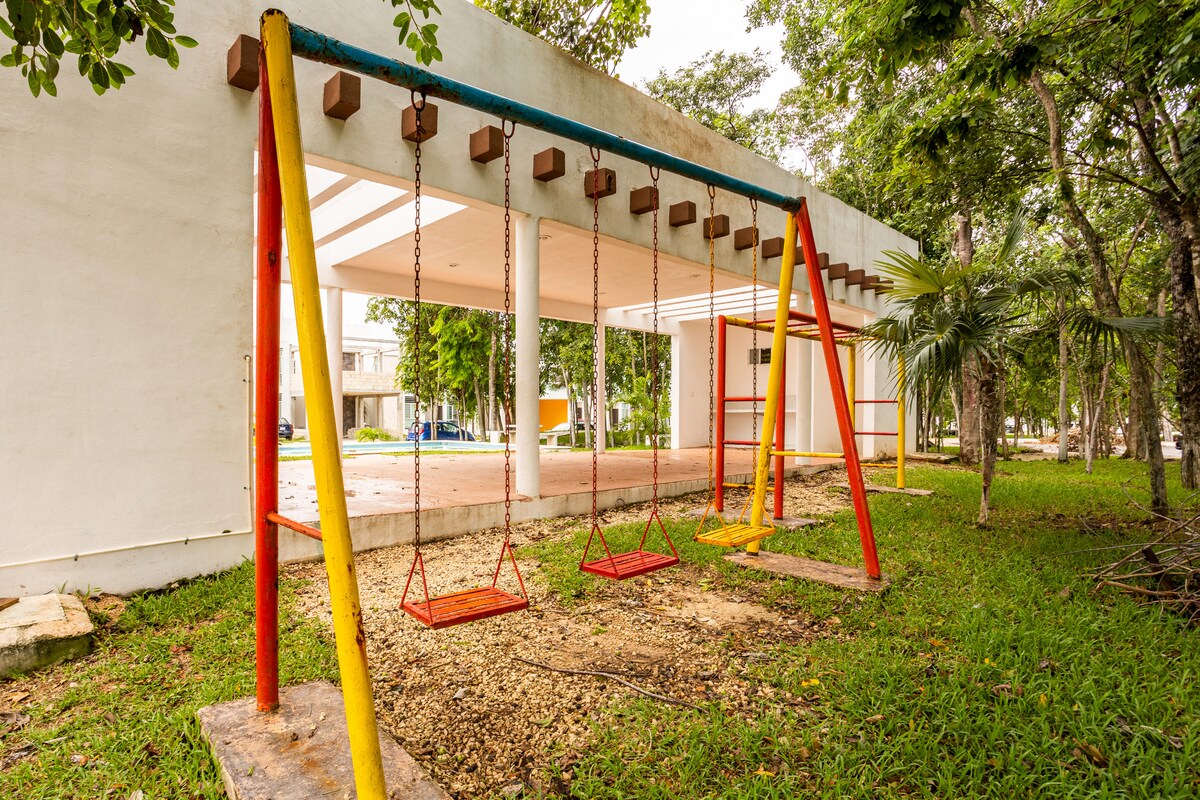 A playground area is visible, featuring two swings with bright red and yellow frames. The ground is covered with gravel and grass. Surrounding trees provide shade, while a spacious building stands in the background, partially obscured by greenery.