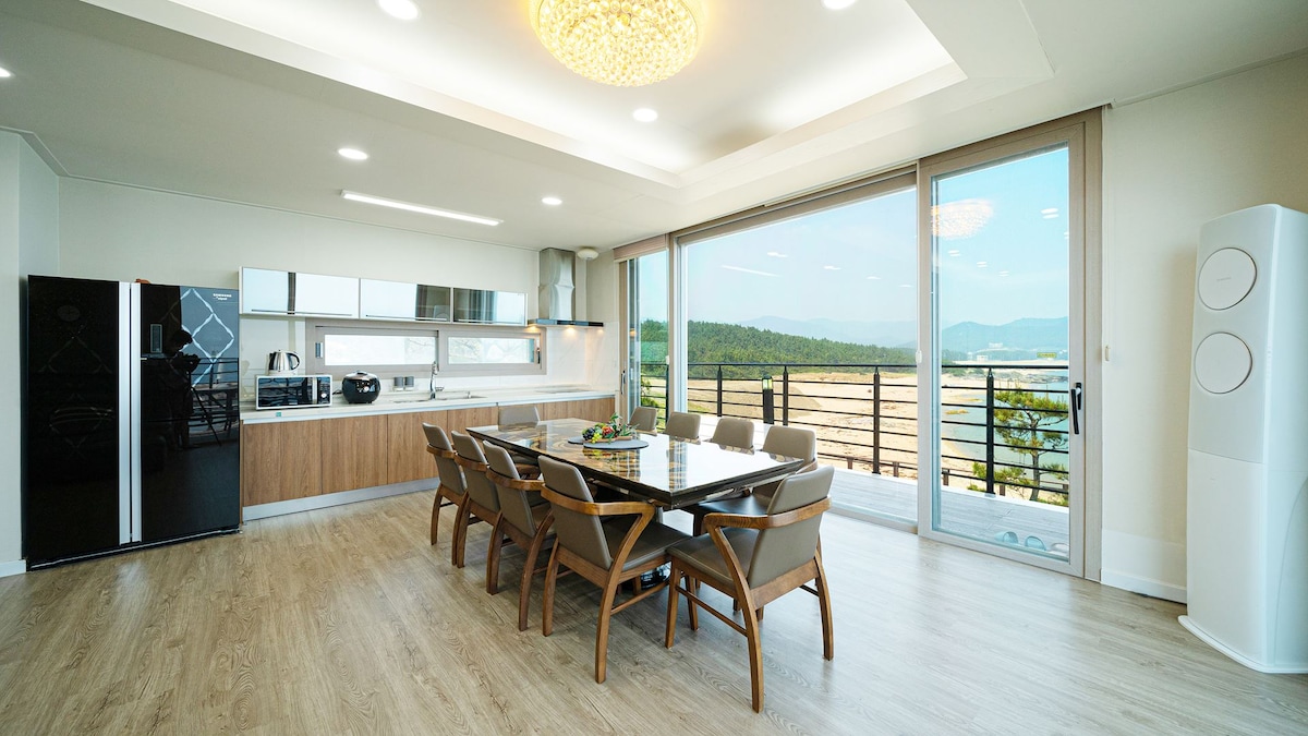 A spacious dining area is furnished with a large table surrounded by ten chairs, complemented by a modern kitchen featuring sleek cabinetry. Expansive glass doors offer a view of the surrounding landscape, allowing natural light to fill the room.