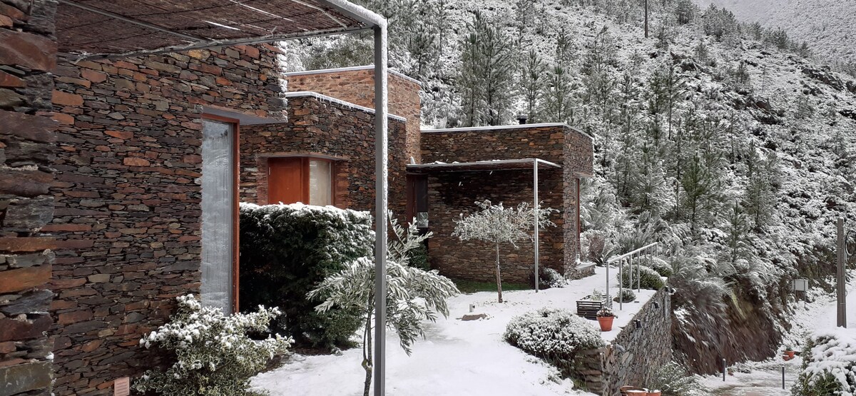 Stone houses are surrounded by a snowy landscape, showcasing a blend of traditional and contemporary architecture. A peaceful garden is visible, with a few scattered plants and trees. The mountains in the background are blanketed in snow, creating a serene atmosphere.