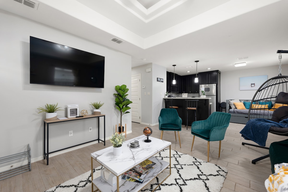 An airy living area features a wall-mounted television and a modern coffee table atop a patterned rug. Stylish green chairs are positioned nearby, while a decorative plant adds a touch of nature. A well-equipped kitchen is visible in the background, blending seamlessly with the space.