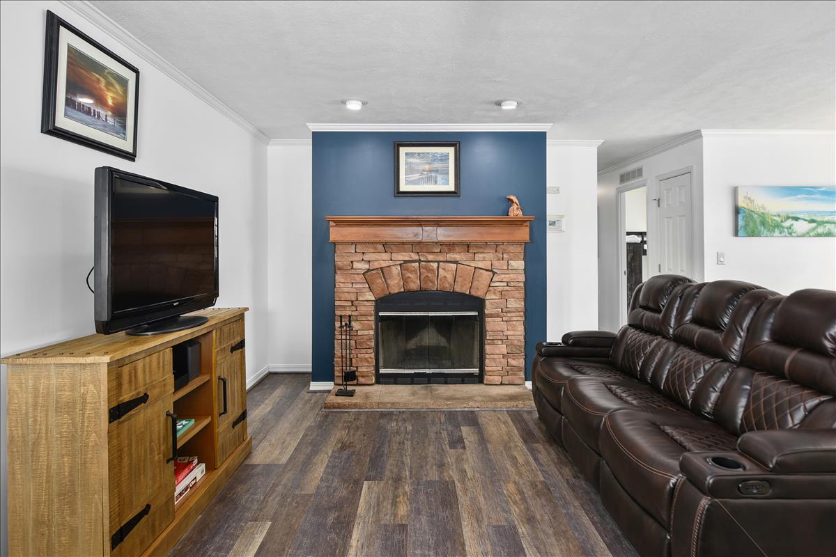 A comfortable living area features a stone fireplace with a wooden mantel as a focal point. A large flat-screen TV is placed on an elegant wooden cabinet. A plush, dark brown sectional sofa offers relaxed seating against a blue accent wall, complemented by framed artwork.