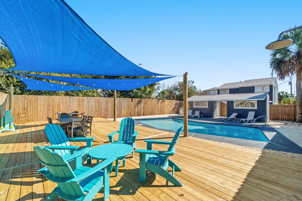A spacious outdoor area is featured with a wooden deck, several bright blue Adirondack chairs surrounding a table, and a large swimming pool. A shade sail provides cover, while a fence offers privacy. The duplex units are visible in the background.