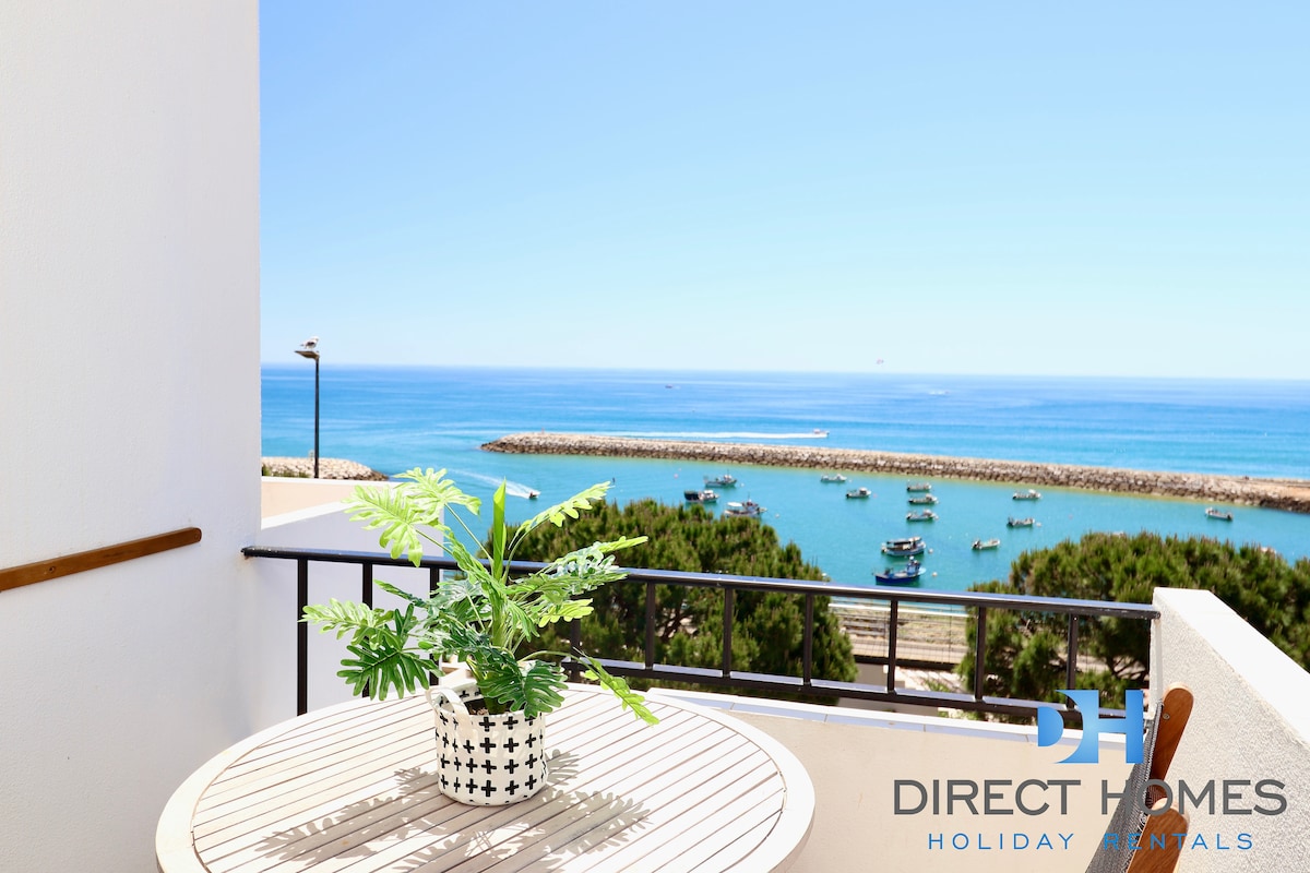 A sunny balcony offers a clear view of the ocean and marina, where fishing boats are visible. A potted plant adds a touch of greenery to the outdoor space, which is surrounded by railings, enhancing the inviting setting for relaxation.