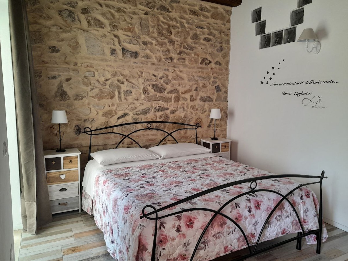 A sleeping area is featured with a spacious bed adorned with floral bedding. Exposed stone walls provide a rustic backdrop, complemented by modern nightstands with lamps. Decorative elements include wall art and framed pictures, enhancing the serene ambiance of the room.