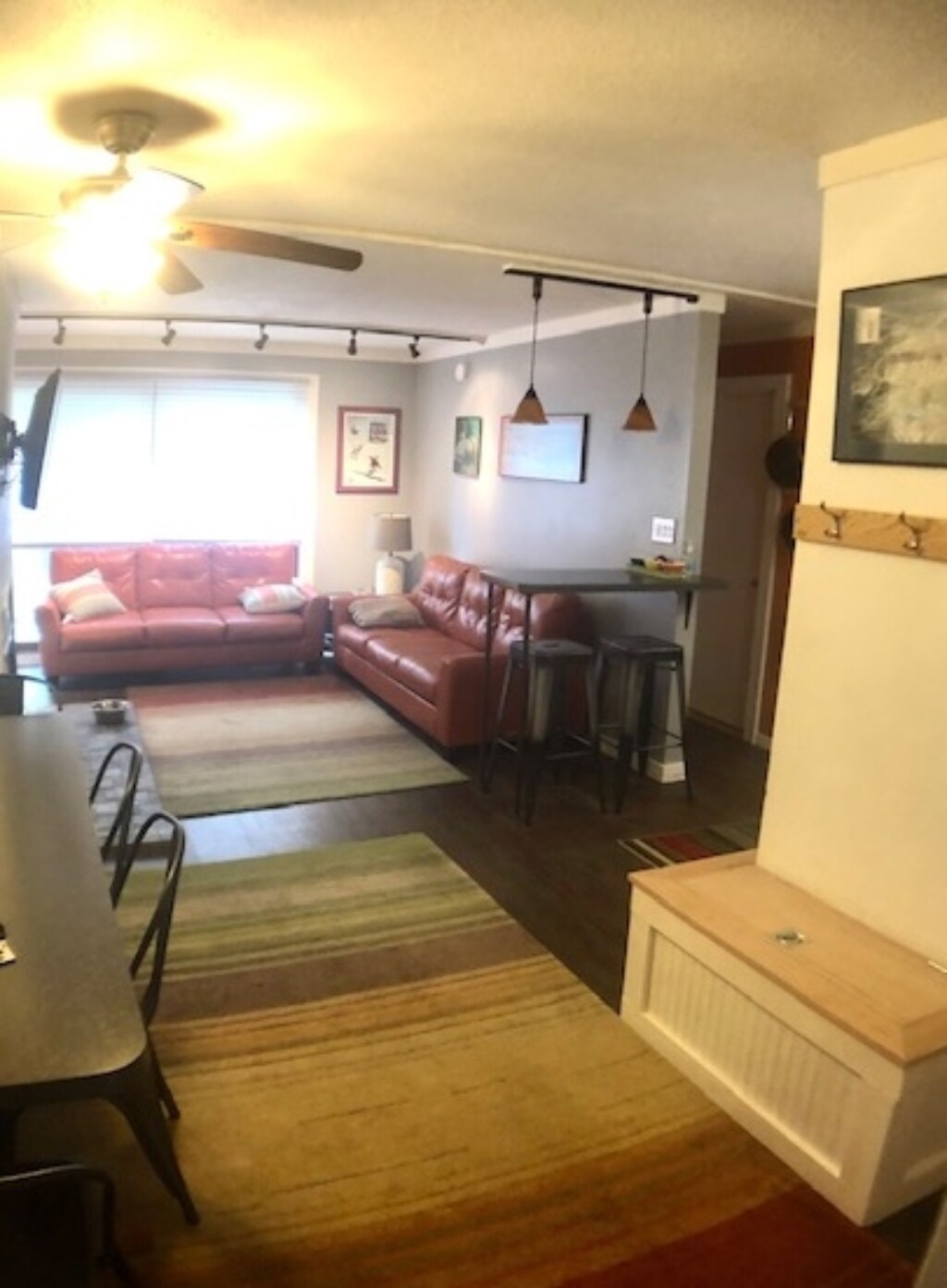 A living area features a red leather sofa and a dining table accompanied by metal chairs. Colorful area rugs are placed on the floor, creating an inviting space. Natural light streams through a window with light curtains, illuminating the room.