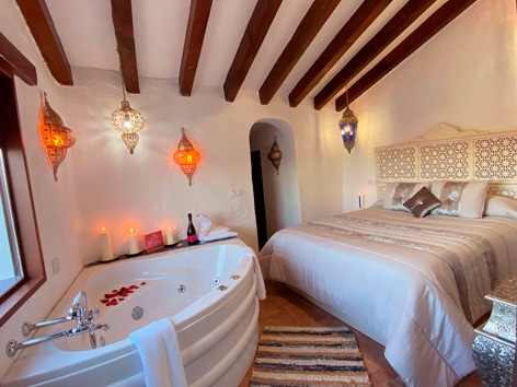 A well-appointed bedroom features a spacious bed with decorative pillows and fabric headboard. A modern jacuzzi is positioned beside the bed, with ambient lighting from hanging lanterns. The warm tones of the wood beams and soft furnishings create a cozy environment.