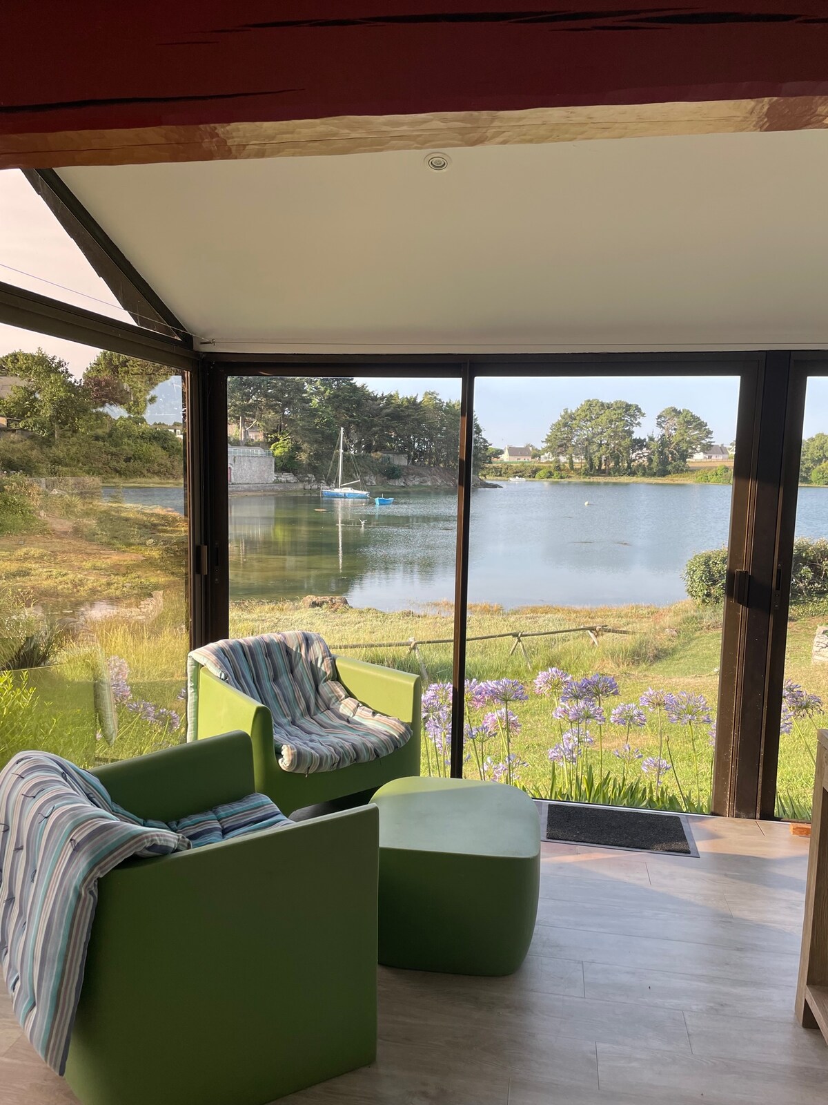 Seating areas with striped cushions are positioned near large glass doors, offering views of the tranquil estuary. Lush greenery and blooming flowers are visible outside, while a sailboat is anchored in the calm waters, enhancing the serene atmosphere.