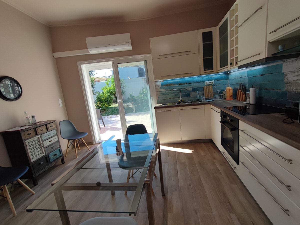 A modern kitchen features sleek cabinetry and a glass dining table surrounded by four chairs. Natural light fills the space from large sliding doors, which open to a garden area. Light wood flooring adds warmth to the room, complemented by a tiled backsplash.