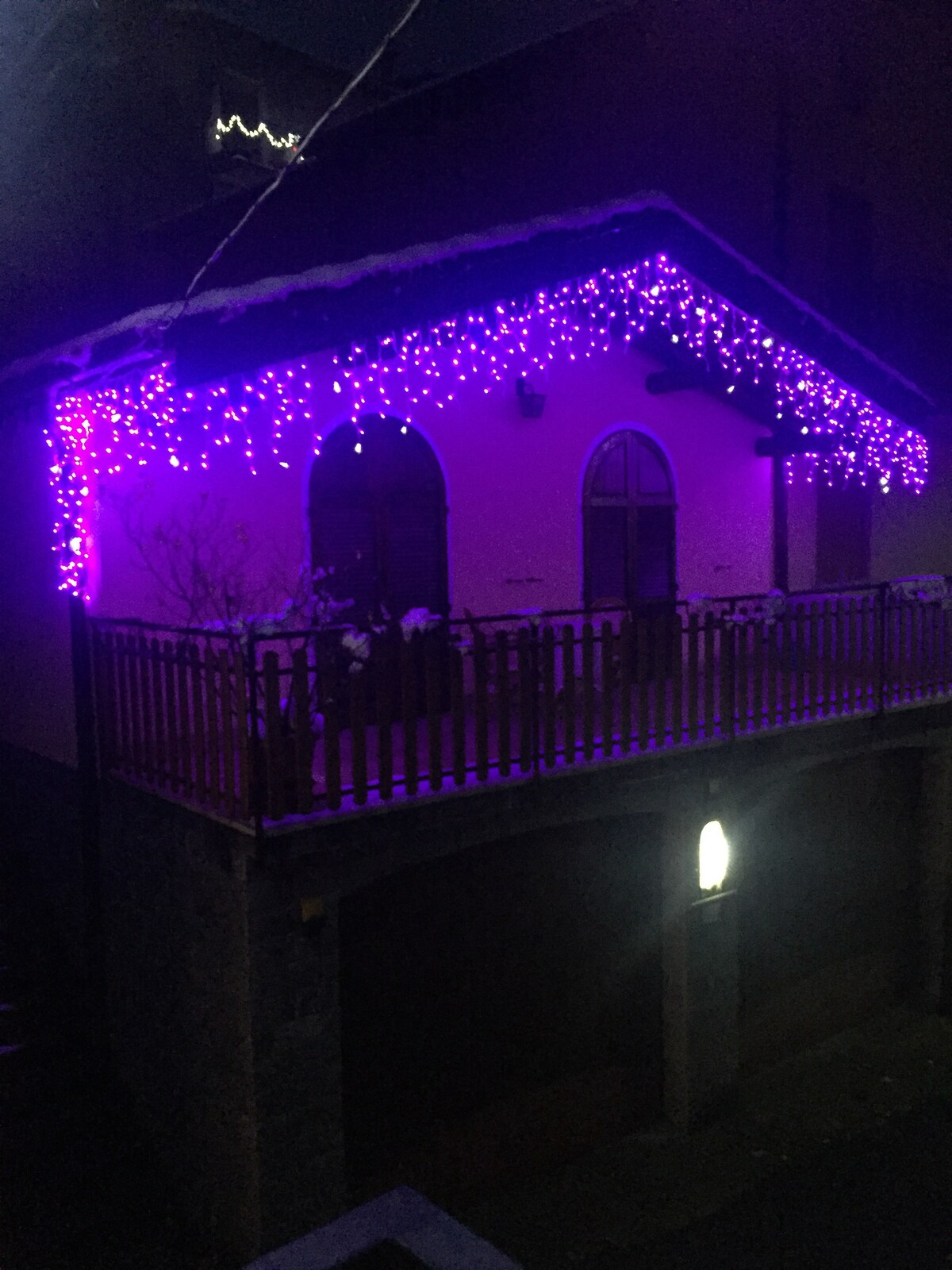 The exterior of the chalet is adorned with purple string lights, illuminating the entrance and balcony area. A wooden railing encompasses the balcony, where snow can be seen on top. The structure's design features arched windows and a roof with a slight overhang.