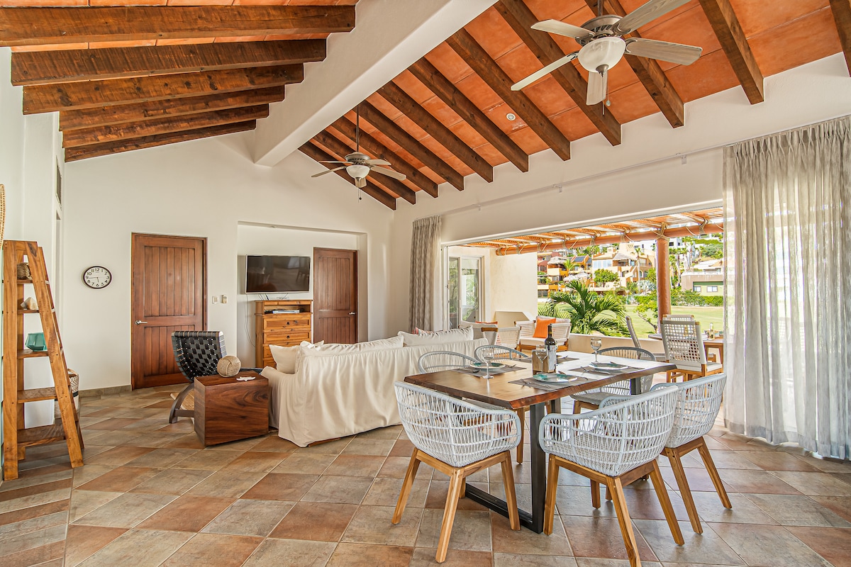 An open-concept living area is featured, characterized by vaulted wooden ceilings and ample natural light. A spacious seating arrangement includes a cozy sectional and a wooden table with woven chairs. Sliding glass doors lead to an outdoor patio, enhancing the indoor-outdoor living experience.