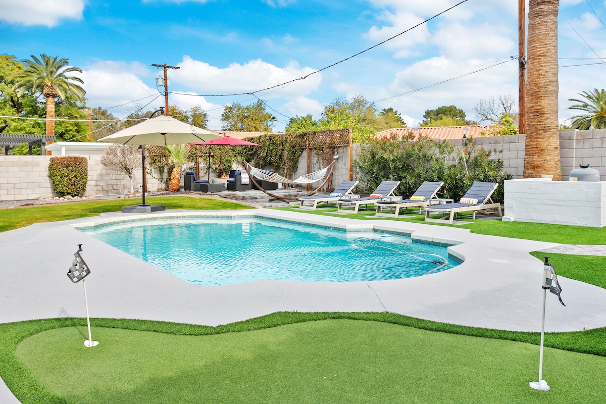 A heated pool is surrounded by a spacious yard, featuring lounge chairs and an umbrella for shade. A putting green adds a recreational element, while palm trees and shrubs provide a lush backdrop. Hammocks are visible, inviting relaxation in the vibrant outdoor space.
