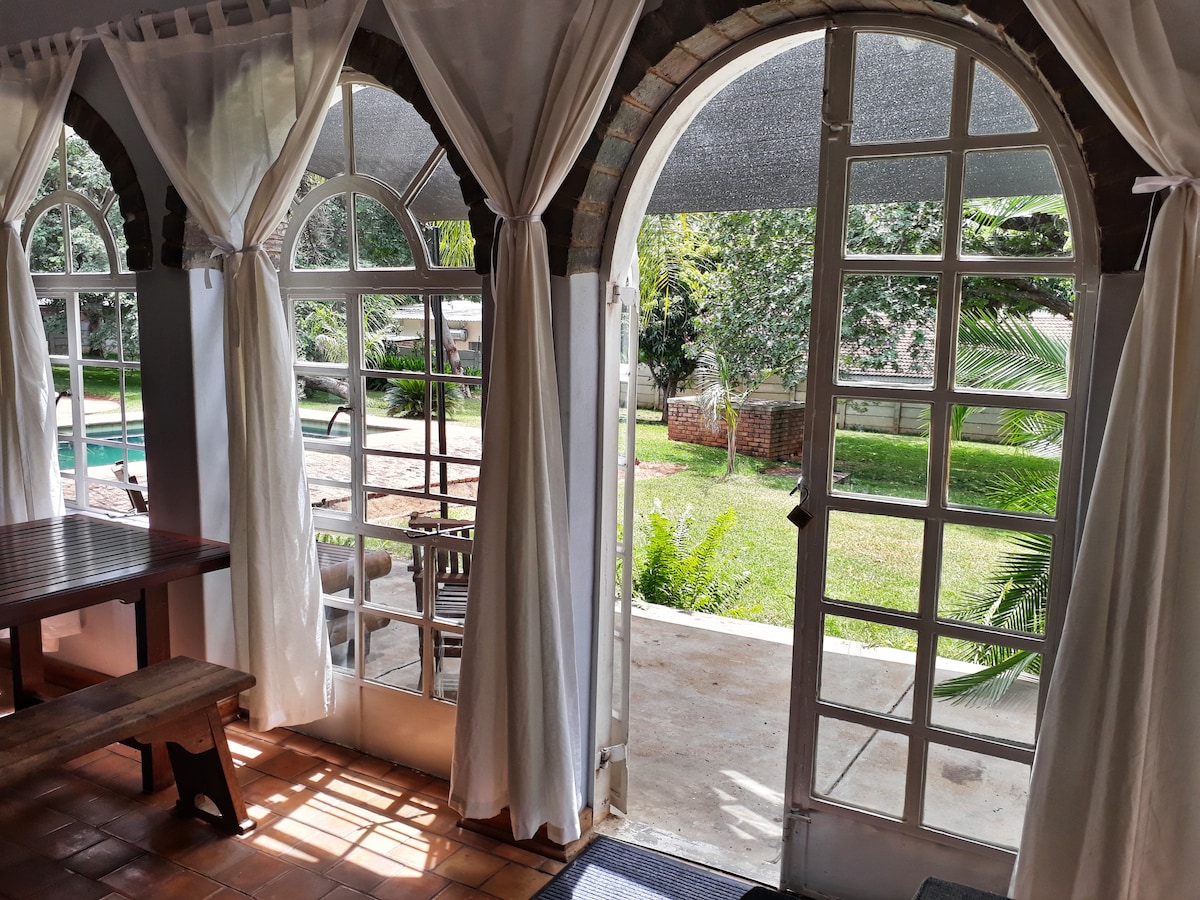 Large arched windows with sheer white curtains frame a view of the tranquil garden and swimming pool. An outdoor patio area is visible, featuring lush greenery. A rustic wooden dining table is positioned indoors, inviting a connection with the natural surroundings.