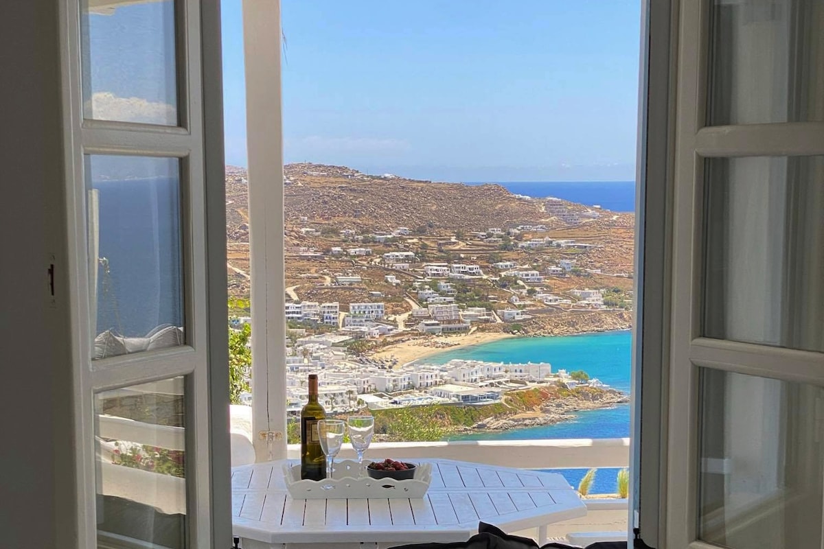 A view from the balcony reveals a scenic landscape, with rolling hills and the azure sea in the distance. A small table is set for two, featuring a bottle of wine and two glasses, inviting relaxation and enjoyment of the surrounding beauty.