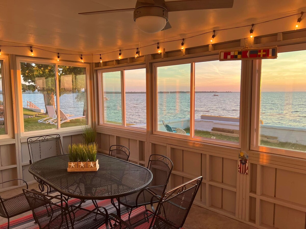 A screened-in porch features a large dining table surrounded by black metal chairs. Large windows offer views of the water, illuminated by soft evening light. String lights are hung along the ceiling, creating a relaxed atmosphere. The porch overlooks the lake, enhancing outdoor dining experiences.