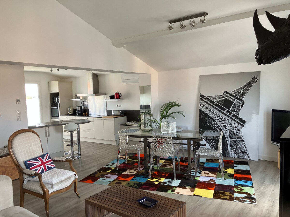 A spacious living area features a vibrant, multi-colored rug underneath a glass dining table surrounded by transparent chairs. A black-and-white artwork depicting the Eiffel Tower takes center stage on one wall, while a modern kitchen with sleek appliances is visible in the background.