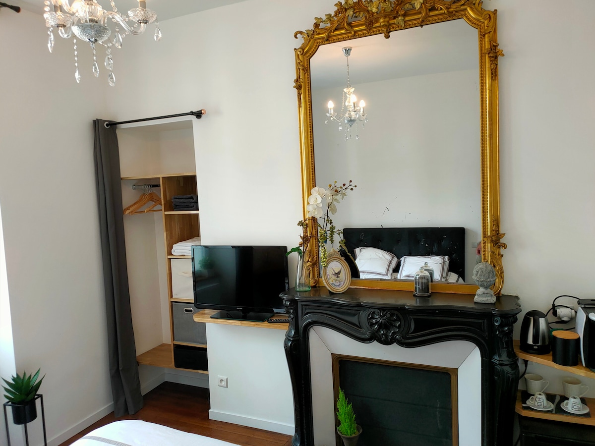 A charming room features a classic marble fireplace and a large, ornate golden mirror above it. A television and a small coffee station are placed nearby. A wardrobe and shelving unit provide storage. Light pours in through the window, enhancing the serene ambiance.