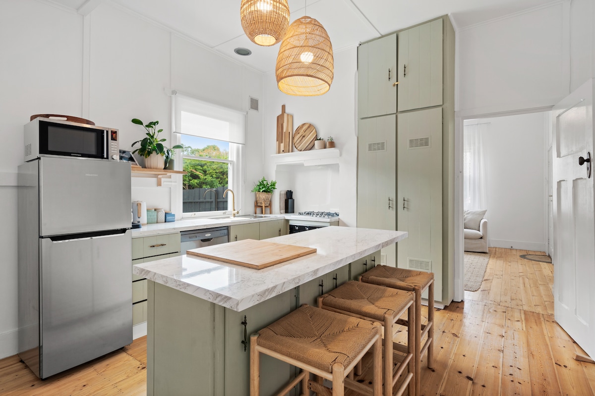 A renovated kitchen features a central island with a marble countertop and four woven stools. Light green cabinetry complements a stainless steel refrigerator and a microwave. Large windows provide natural light, while woven pendant lights hang overhead, adding texture to the airy space.