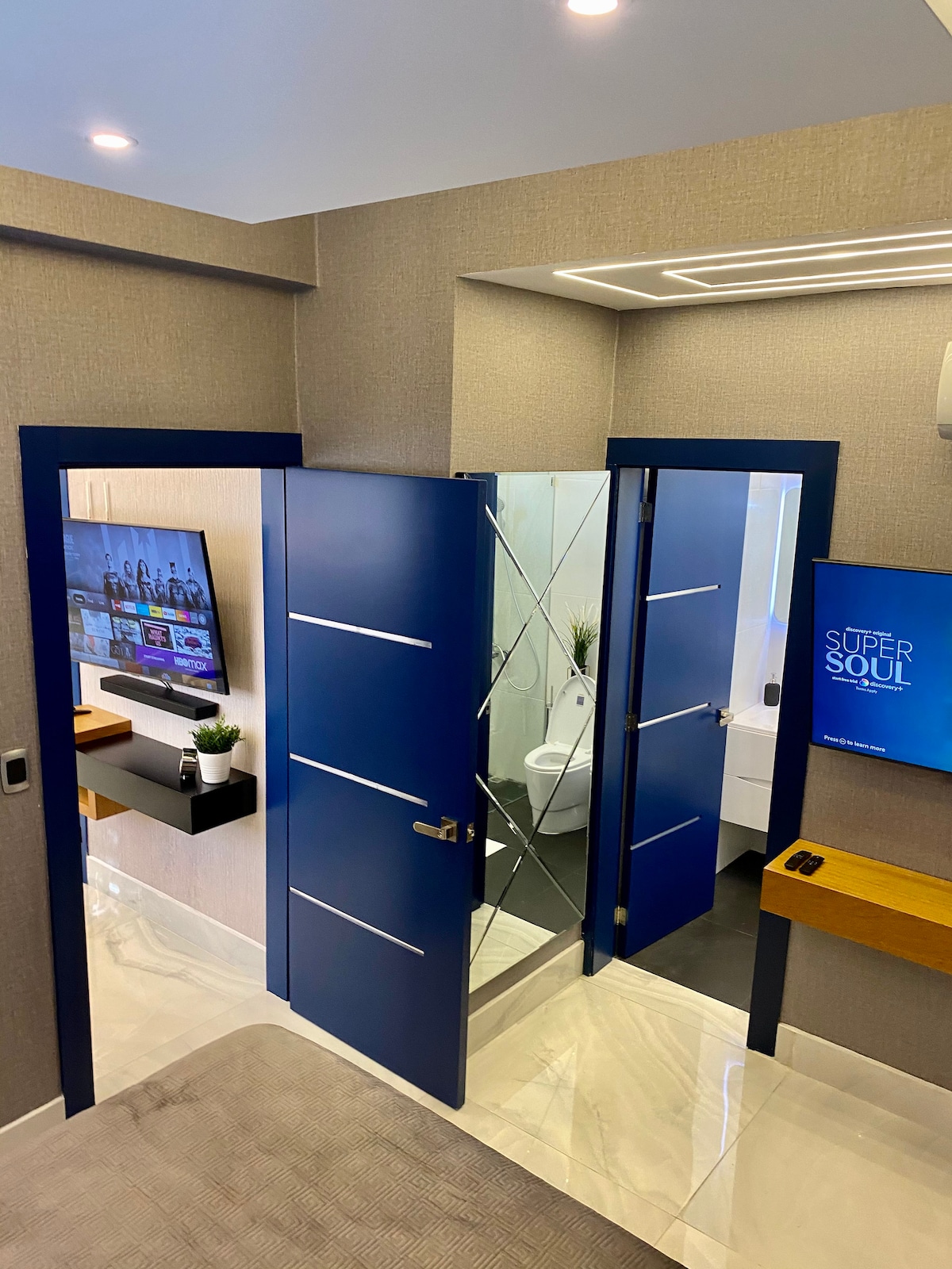 A modern entryway is characterized by sleek blue doors leading to a bathroom and living space. One door opens to reveal a compact restroom with glass panels. A flat-screen TV is mounted on the wall, next to a wood shelf featuring a small plant.