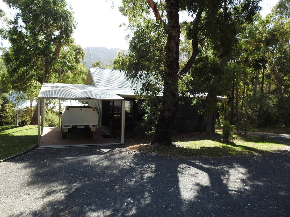 A modern cottage is nestled among lush greenery, featuring a carport that accommodates a vehicle. The structure showcases a combination of metal and wood materials, with large windows allowing natural light to enter. A gravel driveway leads up to the entrance.