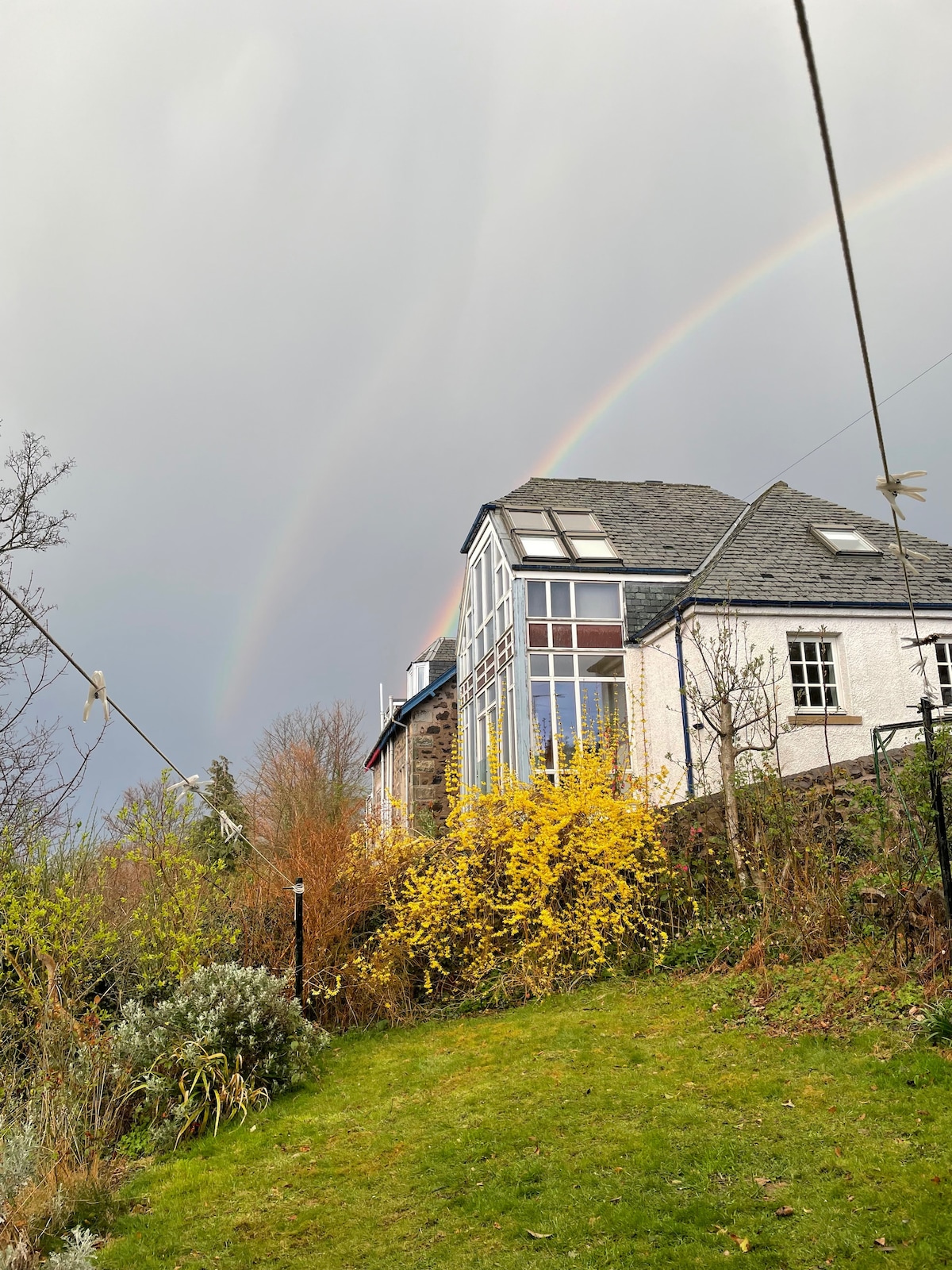 A view of the property is displayed against a moody sky, featuring a double rainbow. The house's large windows reflect the natural surroundings, while vibrant yellow flowers and greenery are visible in the foreground. The landscape suggests a serene setting.