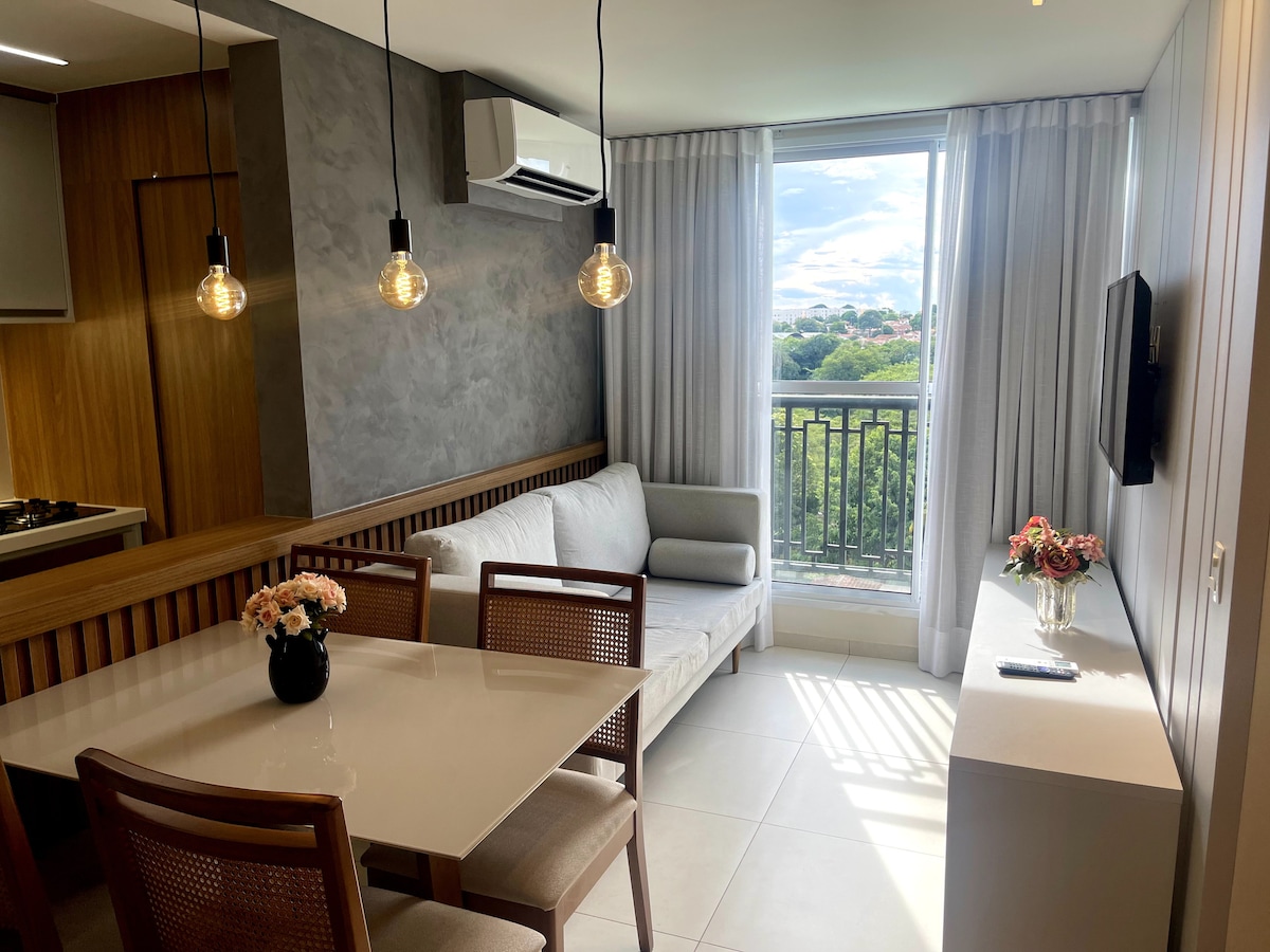 A modern living area features a light-colored sofa accompanied by a stylish side table. A dining table with four wooden chairs is positioned nearby. Large windows provide natural light and views of the surrounding landscape, with pendant lights accentuating the space.