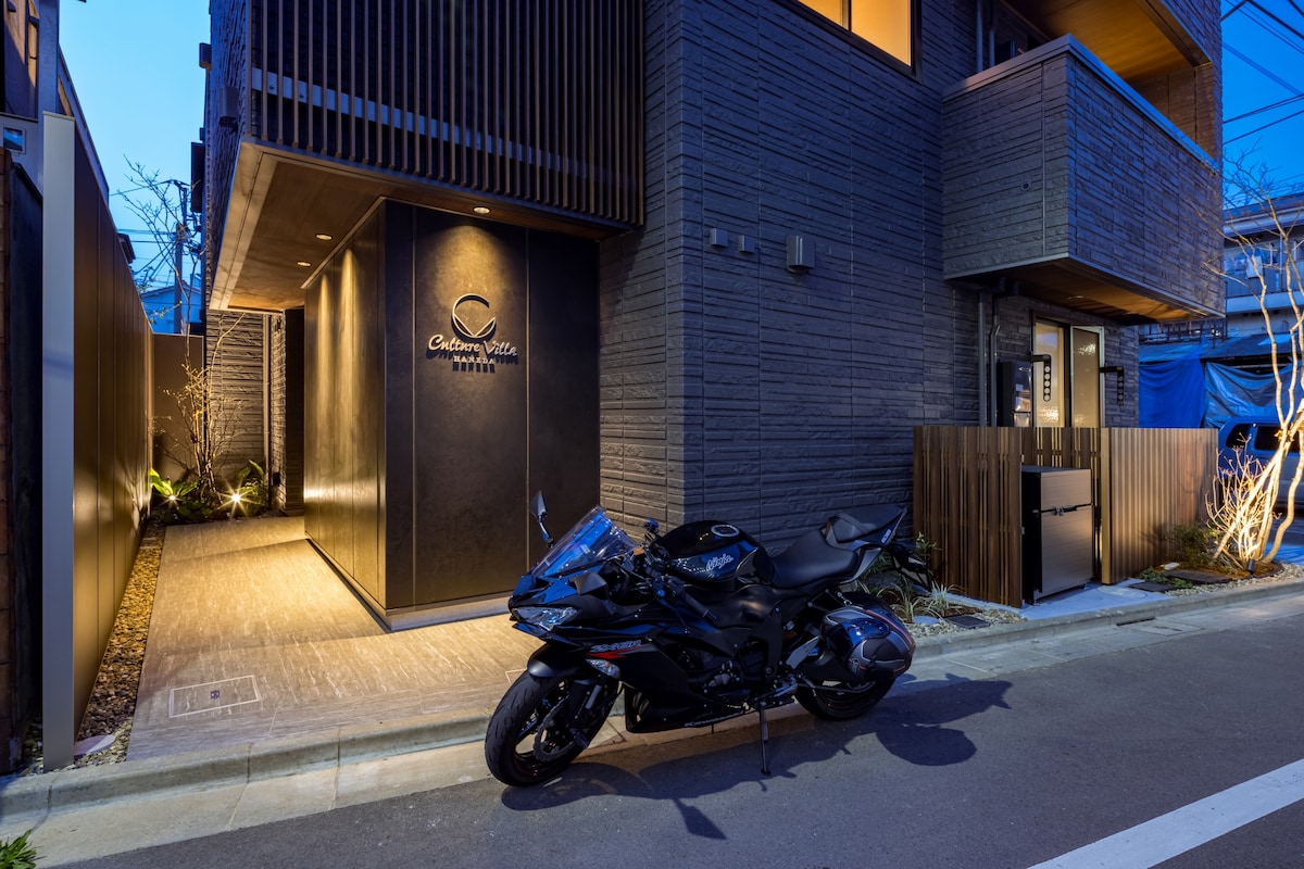 A modern building entrance is visible, featuring a sleek dark façade with an illuminated sign. A motorcycle is parked in front, positioned on a well-maintained pathway lined with subtle lighting. The surrounding area shows minimal landscaping, enhancing the contemporary aesthetic.