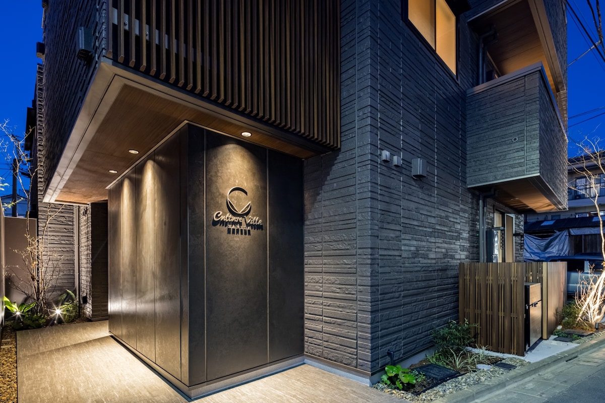 The modern building features a sleek, dark exterior with intricate wooden paneling. The entrance is marked by a stylish logo that highlights the property name. Subtle lighting enhances the architectural details, providing a welcoming feel during dusk.