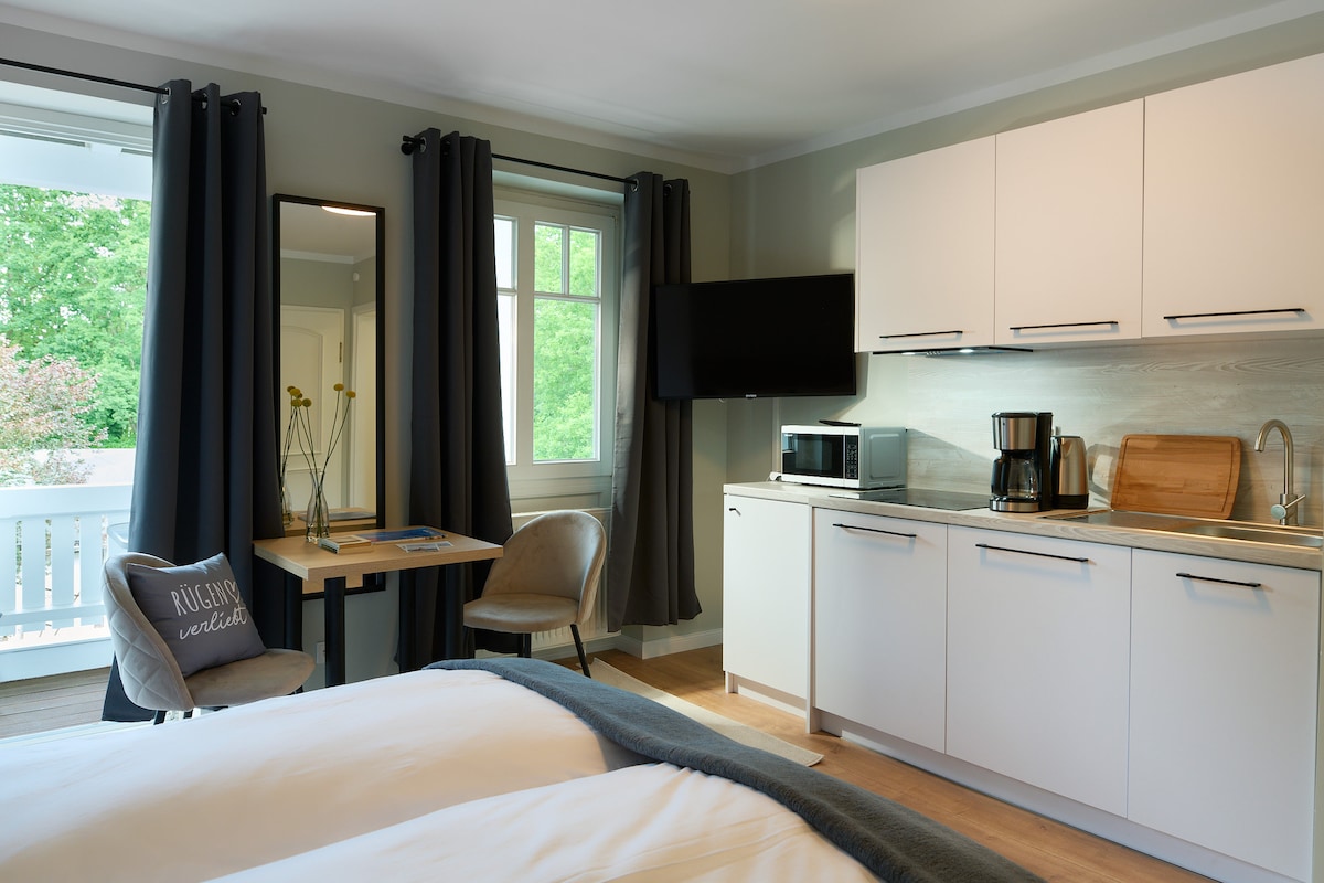The image shows a well-organized studio apartment featuring a spacious sleeping area with two beds covered in soft linens. A modern kitchen area is equipped with essential appliances, while a small dining table with chairs is positioned by a window dressed in dark curtains.
