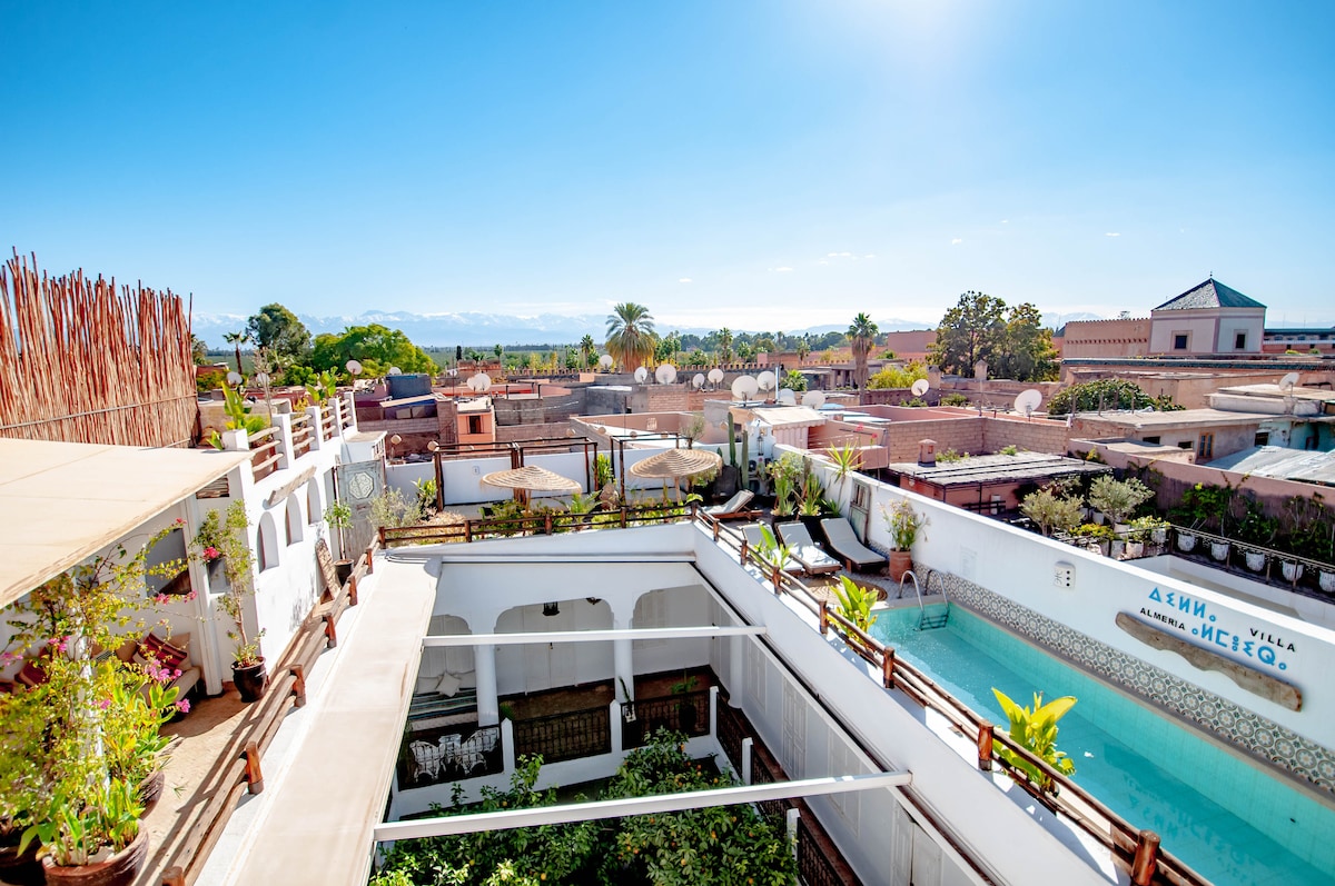 A panoramic view of the rooftop terrace reveals a serene swimming pool and lush greenery. The surrounding area showcases traditional Moroccan architecture, with rooftops and gardens extending towards the distant mountains against a clear blue sky.