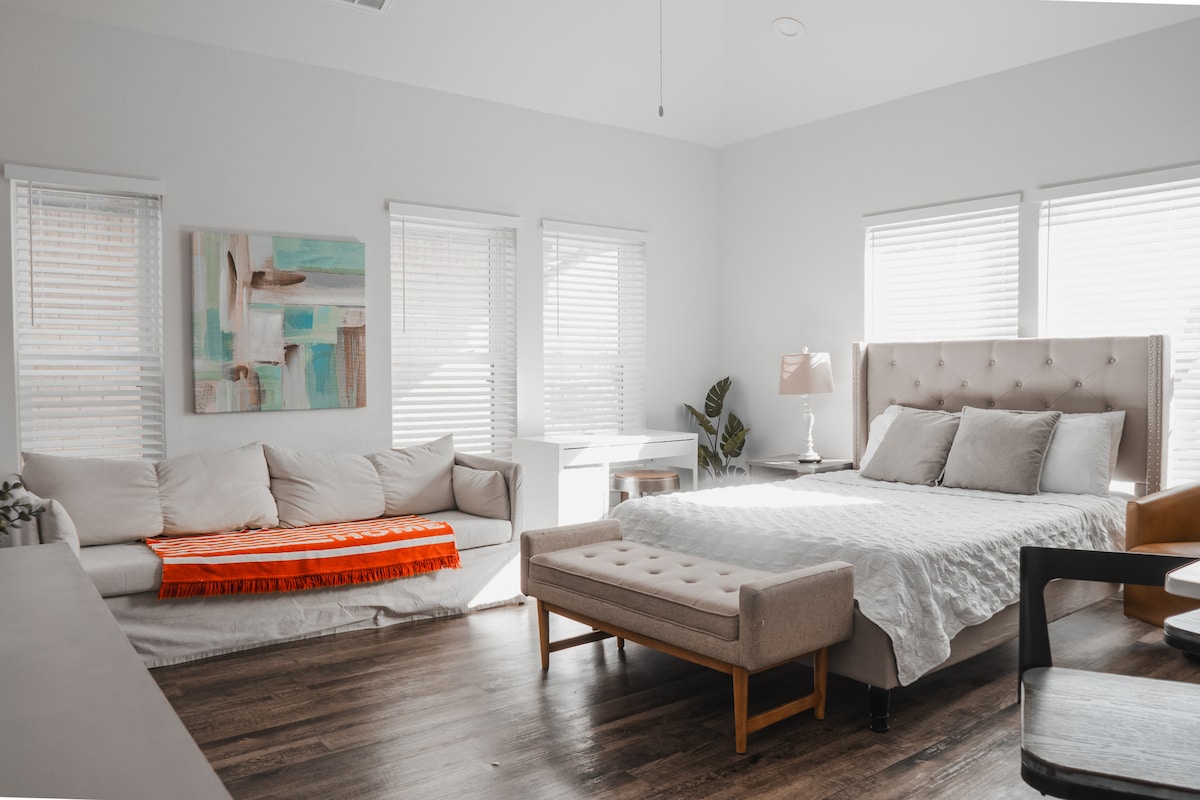 The studio features a spacious layout with a queen size bed dressed in soft linens. A light-colored sofa adorned with an orange throw complements the decor. Natural light pours through multiple windows, enhancing the calming atmosphere of the contemporary space.