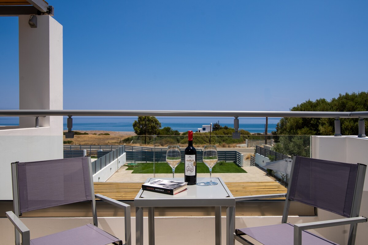 A charming outdoor balcony features two lightweight chairs positioned around a small table. A bottle of wine and two glasses rest on the table, with a scenic view of the sea and surrounding landscape visible in the background.