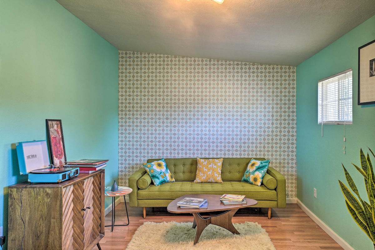 A cozy living area features a green sofa adorned with colorful cushions. The walls are painted in a light turquoise hue, complemented by a patterned accent wall. A wooden coffee table is centered in the space, and natural light enters through a small window.