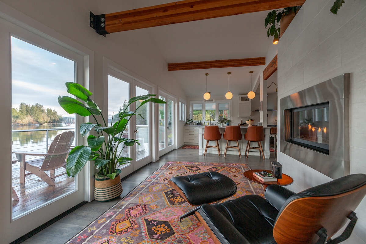 A cozy living area features a sleek chair, a vibrant area rug, and large windows providing views of the water. A plant adds a touch of greenery, while a gas fireplace is set against a neutral wall, creating a relaxing ambience.