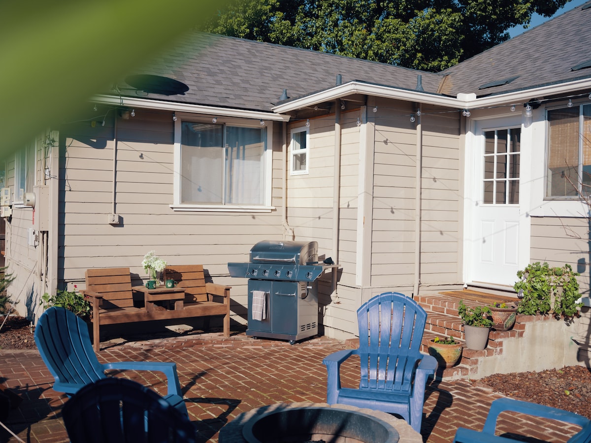 The outdoor patio features a brick surface, accented by blue wooden chairs arranged around a fire pit. A sectional bench offers additional seating near a barbecue grill, and potted plants are visible near the entrance of the cottage.