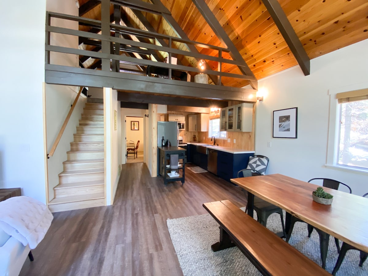 An open-concept living space features high, vaulted ceilings with wooden beams. A staircase leads to the loft. A dining table invites group meals, while a kitchen with modern appliances is visible in the background, complemented by natural light streaming through large windows.