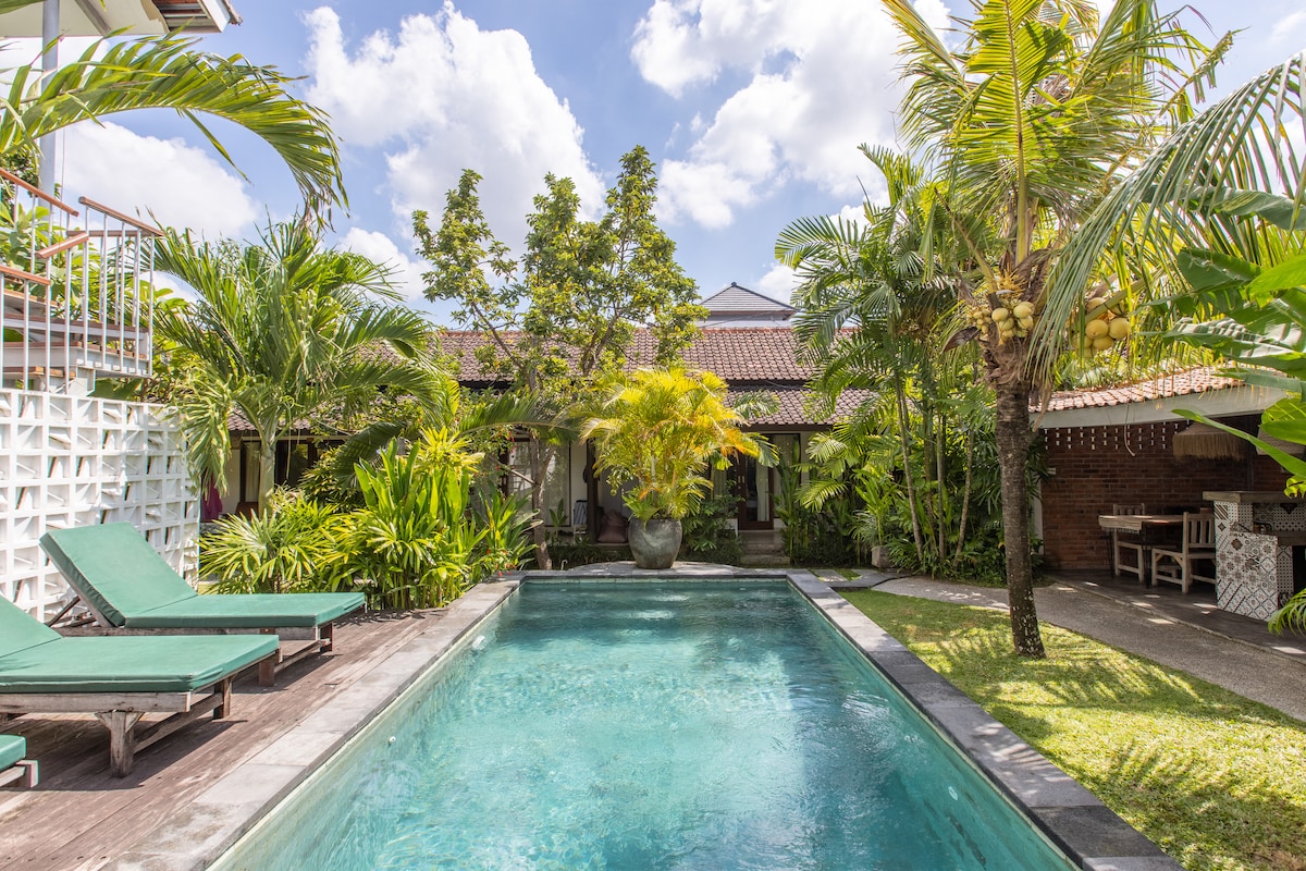 An outdoor area features a swimming pool surrounded by lush greenery and tropical plants. Sun loungers with green cushions are placed beside the pool. The serene setting is enhanced by the clear blue sky, providing a relaxing atmosphere.