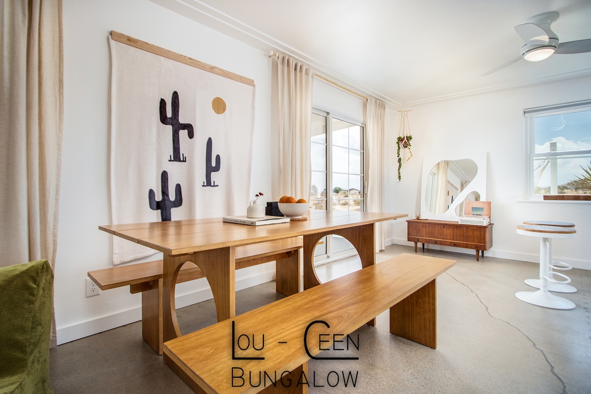 A modern dining area features a wooden table and bench, complemented by a sculptural chair. Natural light fills the space, highlighting a cacti-themed wall hanging. A stylish mirror and a wooden sideboard are seen in the background, adding character to the room.
